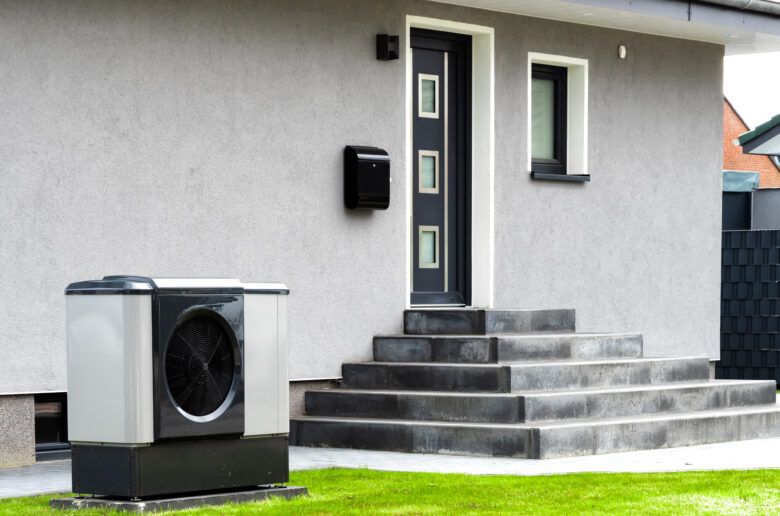 Eine moderne Luft-Wasser-Wärmepumpe steht vor einem grauen Einfamilienhaus. Das Außengerät der Wärmepumpe befindet sich auf einem stabilen Fundament, während im Hintergrund der Hauseingang mit einer dunklen Tür und einer Treppe aus Beton zu sehen ist. Die Umgebung ist gepflegt, mit grünem Rasen vor der Wärmepumpe.