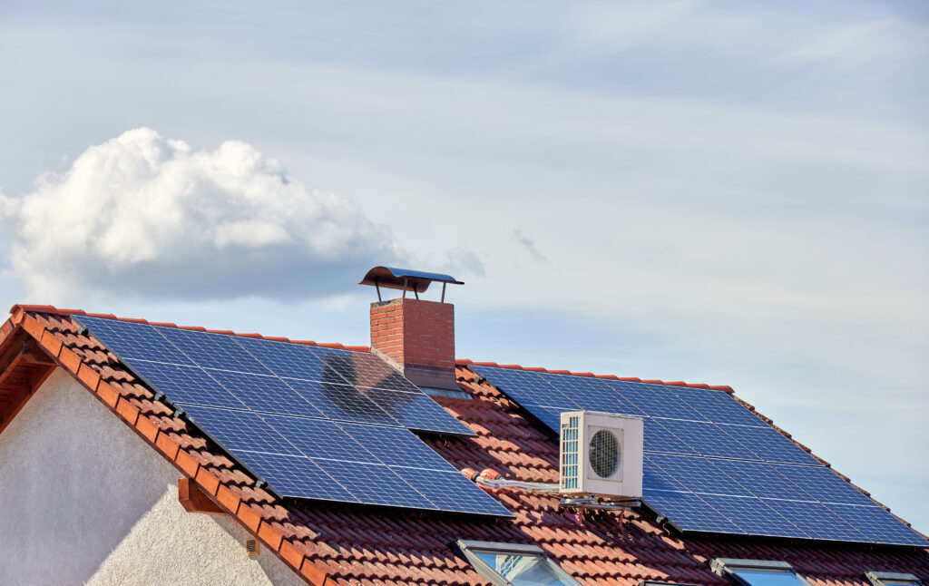 Ein Hausdach mit roten Ziegeln ist mit großen Solarpanelen ausgestattet, die das Sonnenlicht einfangen. Auf dem Dach befindet sich zudem ein Außengerät einer Wärmepumpe.. 