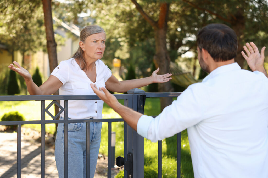 Eine Frau und ein Mann stehen an einem Gartenzaun und gestikulieren miteinander, offensichtlich in einer hitzigen Diskussion. Die Frau schaut verärgert, hebt die Arme und scheint etwas zu erklären, während der Mann mit dem Rücken zur Kamera ebenfalls gestikulierend antwortet. Im Hintergrund sind grüne Bäume und ein gepflegter Garten zu sehen.