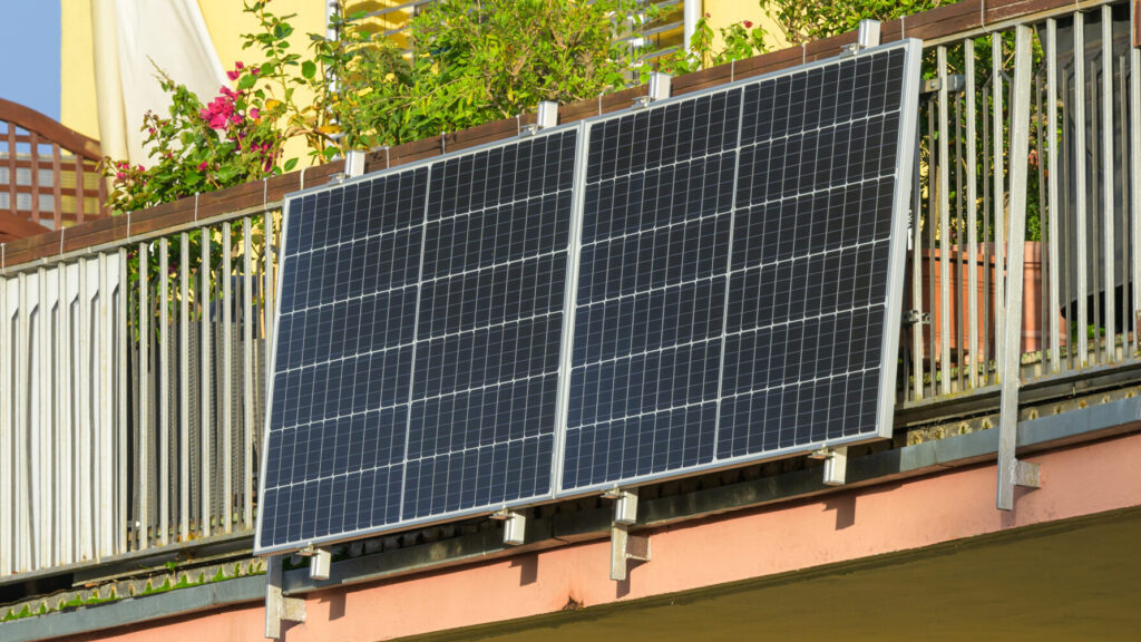 Ein modernes Balkonkraftwerk mit zwei Solarpanels ist an einem Balkon befestigt. Der Balkon ist mit Pflanzen und Blumenkästen dekoriert, während im Hintergrund ein gelbes Wohngebäude sichtbar ist.