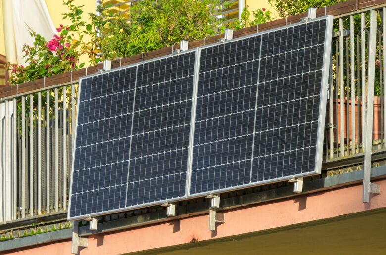 Ein modernes Balkonkraftwerk mit zwei Solarpanels ist an einem Balkon befestigt. Der Balkon ist mit Pflanzen und Blumenkästen dekoriert, während im Hintergrund ein gelbes Wohngebäude sichtbar ist.