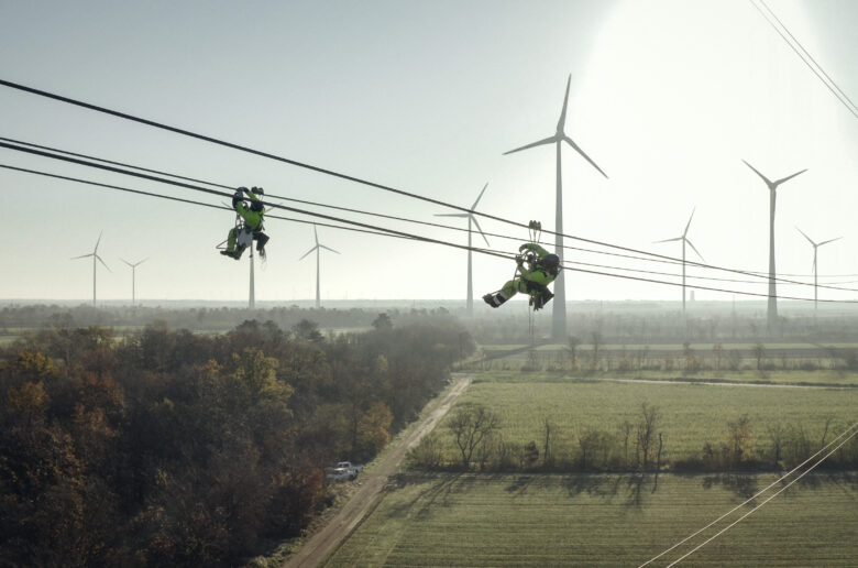 Stromkosten: Zwei Monteure arbeiten an einer Hochspannungsleitung über einer Landschaft mit Windkraftanlagen.